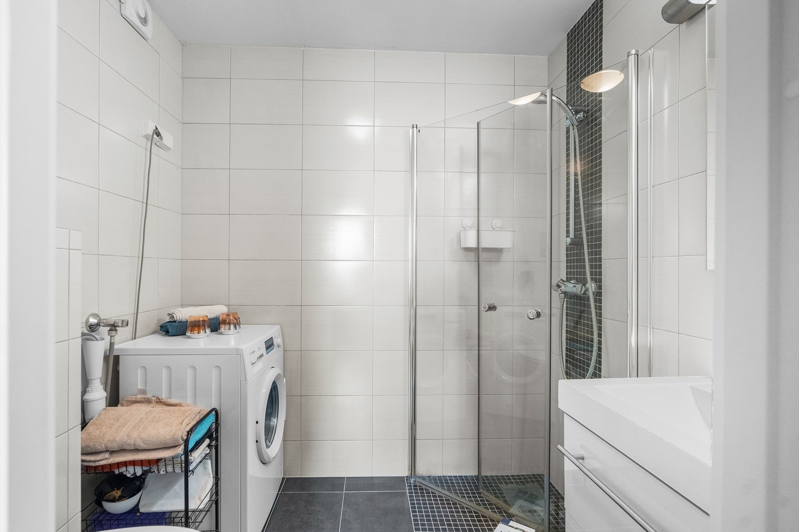 Badet har flislagte overflater, elektrisk avtrekk, servant, veggehengt toalett og dusjhjørne. Det er opplegg for vaskemaskin her og elektrisk avtrekk i rommet. Galleribilde