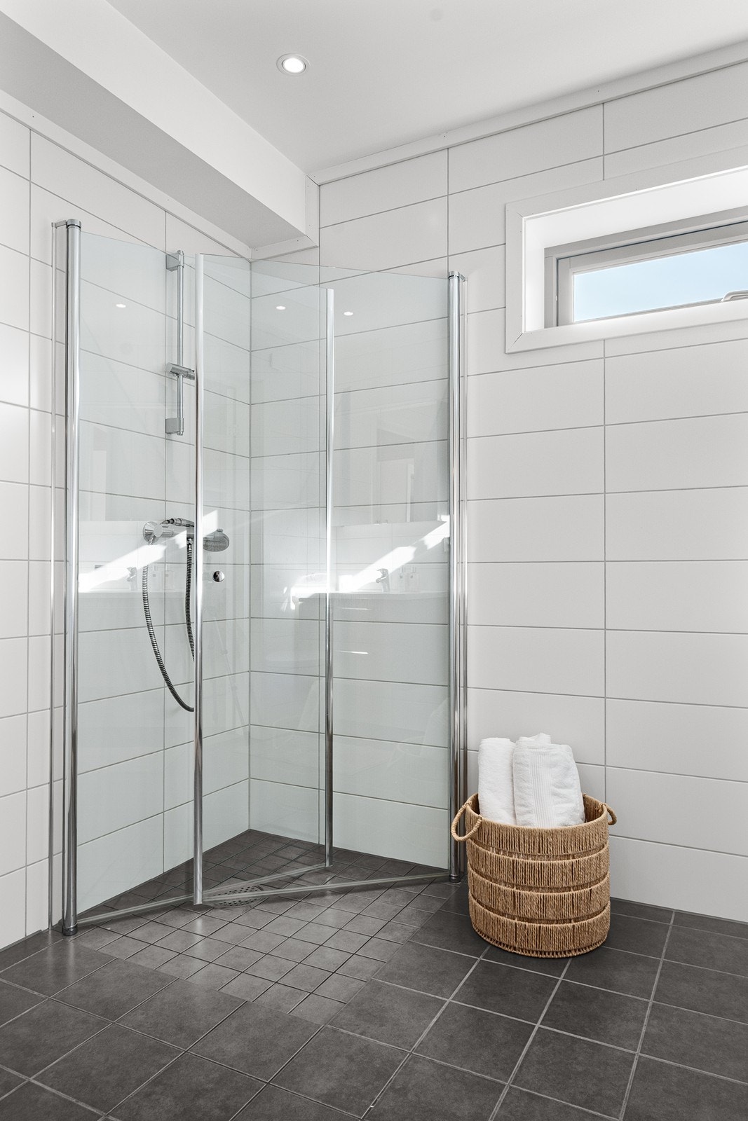 Badet har flislagte overflater, gulvvarme og downlights. Innredningen består av servantskap med skuffeseksjon, veggmontert toalett og dusjhjørne med innfellbare glassdører. Galleribilde