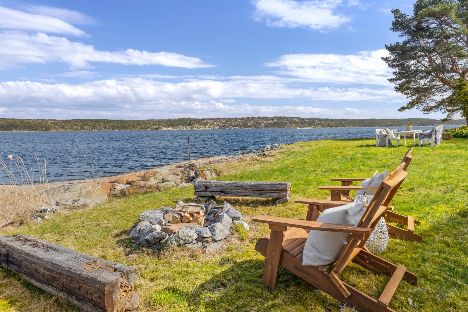 Utsikt over vannet med sitteplasser og bålplass på gressplen. Galleribilde