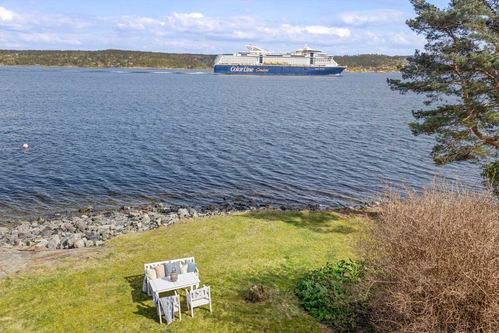 Utsikt mot sjøen med et cruiseskip i bakgrunnen og en liten sittegruppe på gressplen. Galleribilde