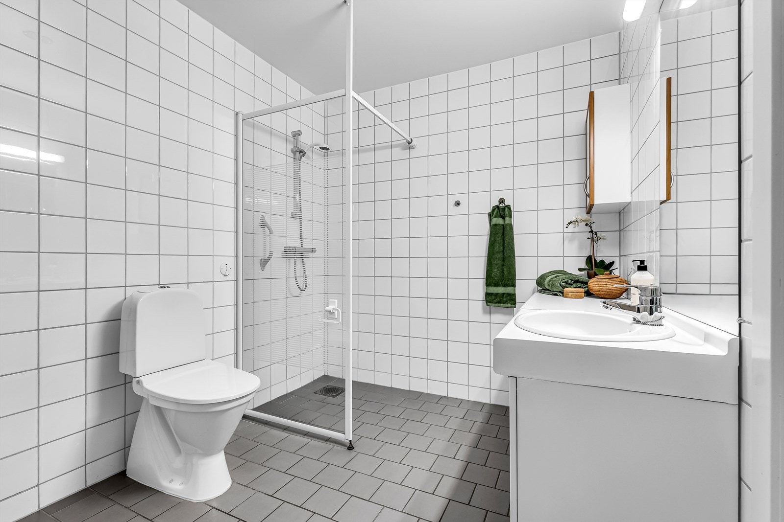 Badet er innredet med servant i møblement med benkeplate og underskap, speil med integrert belysning, veggskap, dusjhjørne og toalett. Galleribilde