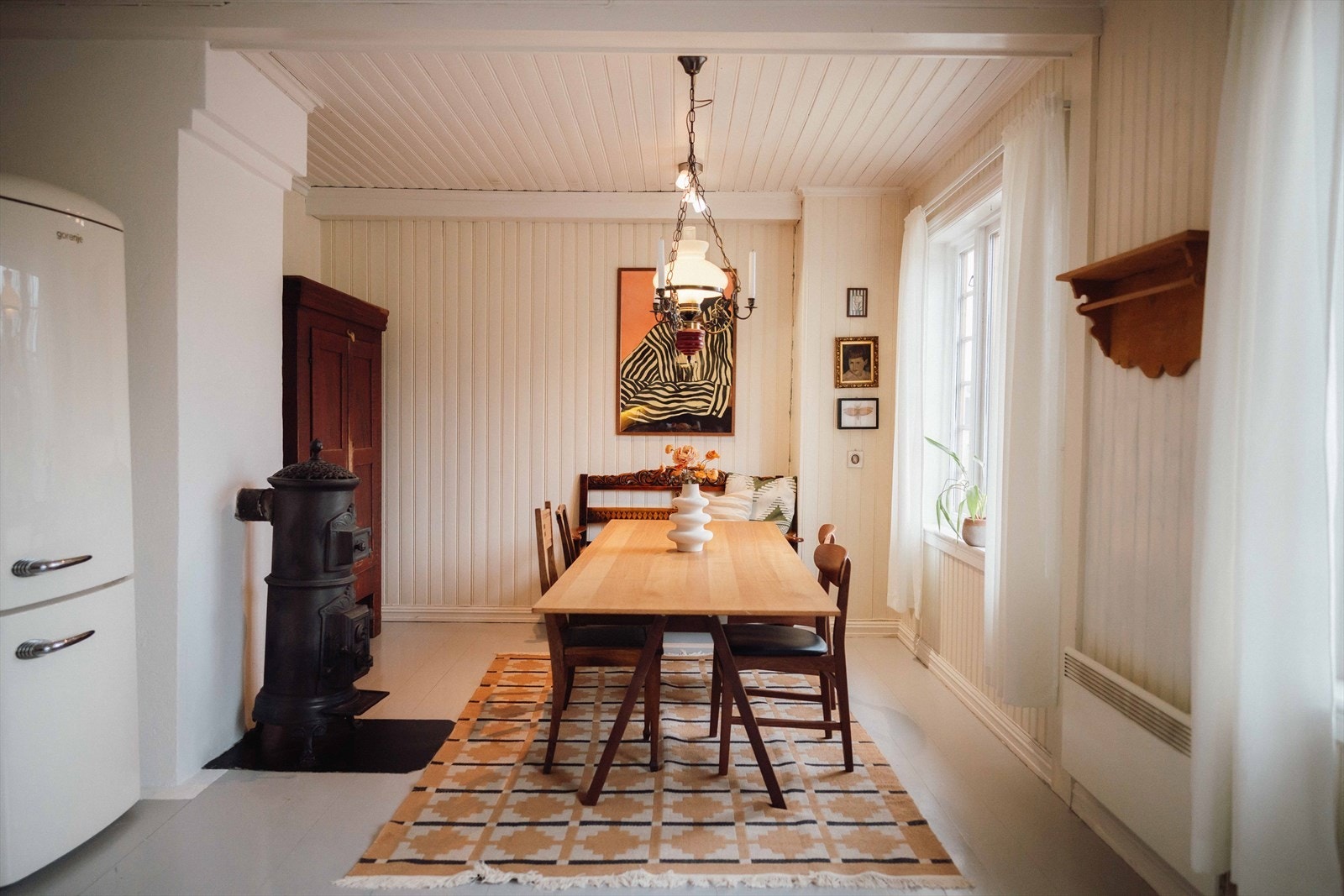 Veggene har stående panel malt i en lys pudderfarge, gulvet er gråmalt og foran en murt pipe er det en eldre etasjeovn. Galleribilde