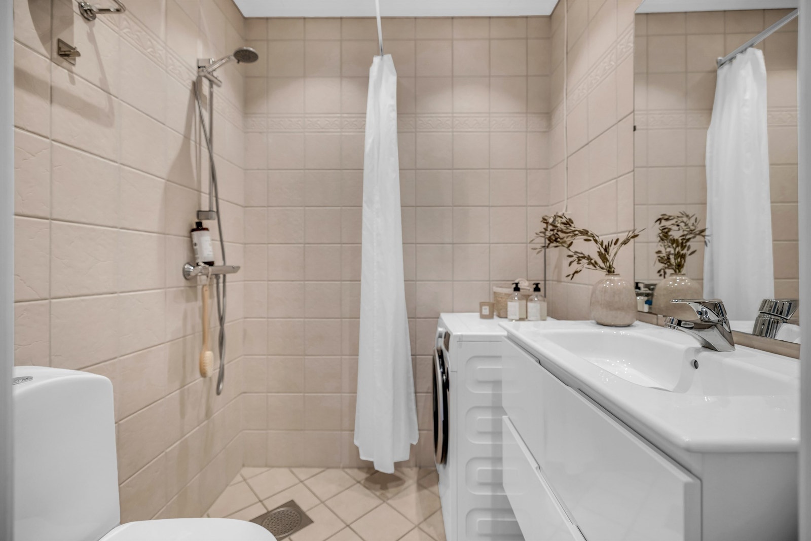 Flislagt bad med varmekabler i gulv, innredet med gulvstående toalett og dusjhjørne som er avgrenset med forheng. Opplegg for vaskemaskin. Galleribilde