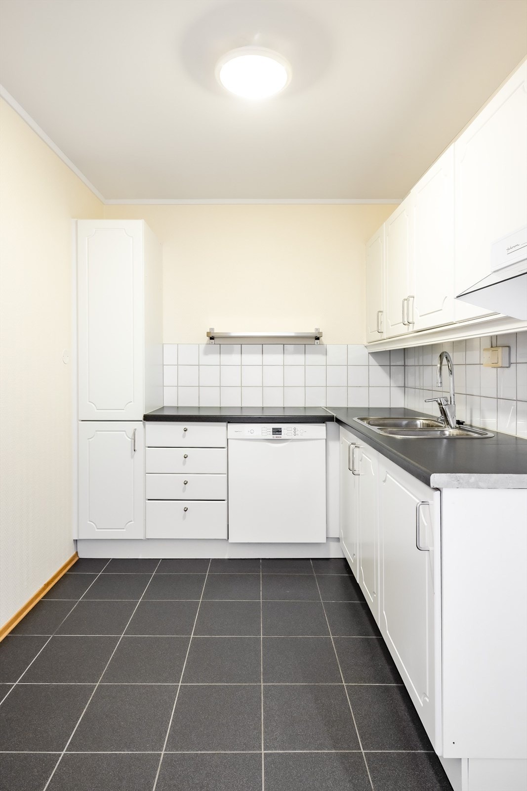 Over- og underskap med hvite, profilerte fronter. Laminert benkeplate med dobbel oppvaskkum i rustfritt stål. I benkeryggen er det fliser. Galleribilde