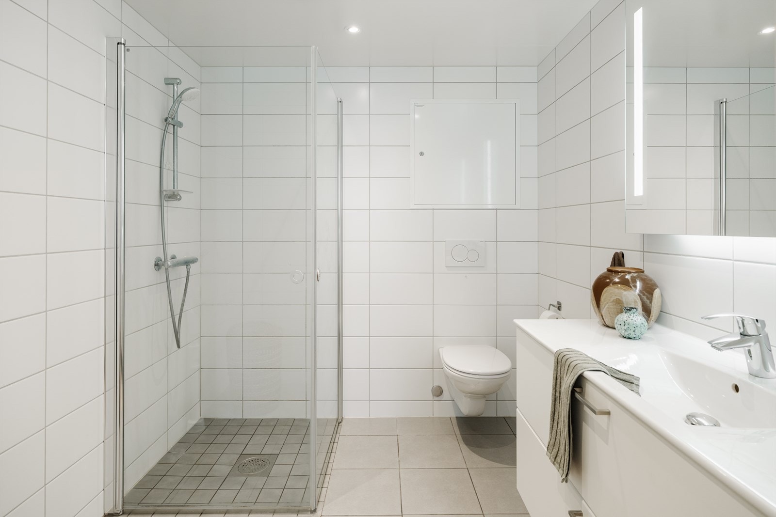 Badet er utstyrt med dusj på gulv med dusjdører i herdet glass, servant med skapinnredning samt vegghengt toalett med innebygget sisterne. Galleribilde
