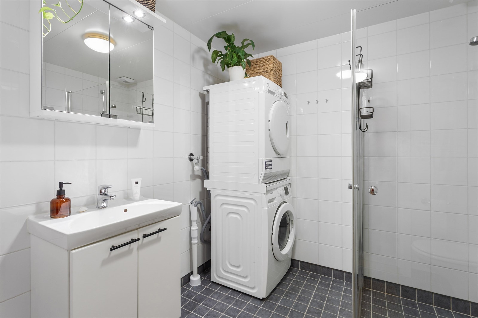 Romslig baderom med hvite fliser på vegg og grå fliser på gulv med varmekabler. Opplegg for både vaskemaskin og tørketrommel. Galleribilde