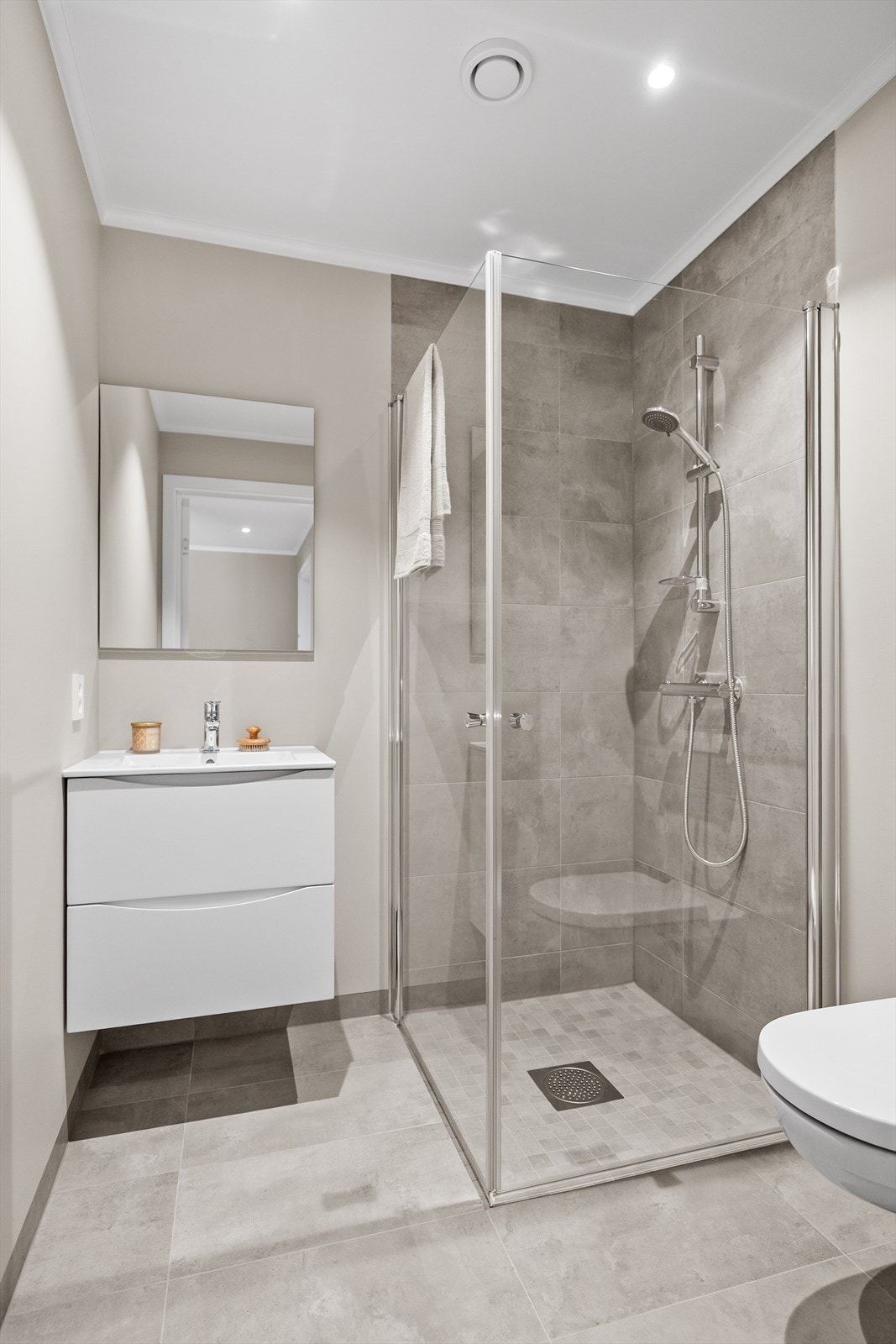 Badet har inngang fra entré og er i delikat utførelse. Utstyrt med hvit innredning med overliggende servant, vegghengt speil, dusjhjørne med innfellbare dører og vegghengt toalett Galleribilde