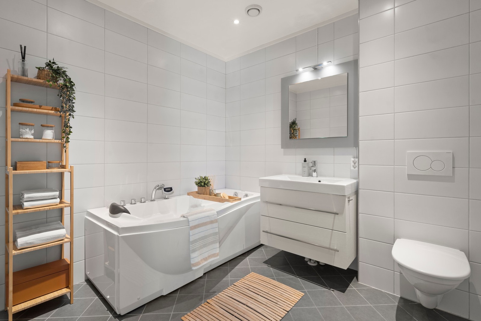 Badet i 1. etasje er romslig og delikat utformet med flislagte gulv og vegger som gir et stilrent og tidløst uttrykk. Her finner du varmekabler i gulvet, badekar, vegghengt toalett og en praktisk servantinnredning som skaper både komfort og gode hverdagsl Galleribilde