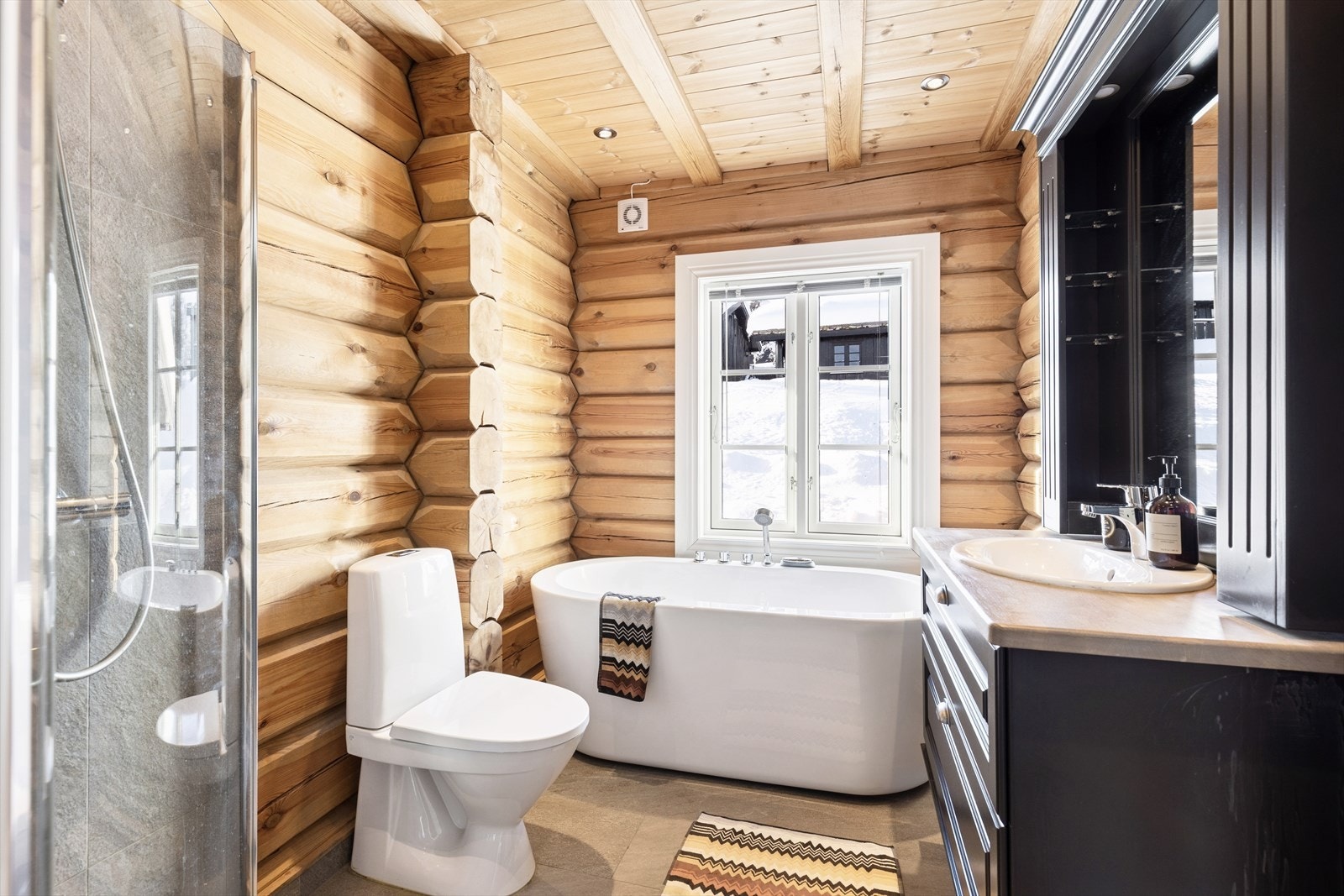 Delikat bad med tømmervegger, fliser på vegg i våtsone og fliser på gulv med gulvvarme. I himling er det trepanel med kunstige bjelker og downlights. Galleribilde