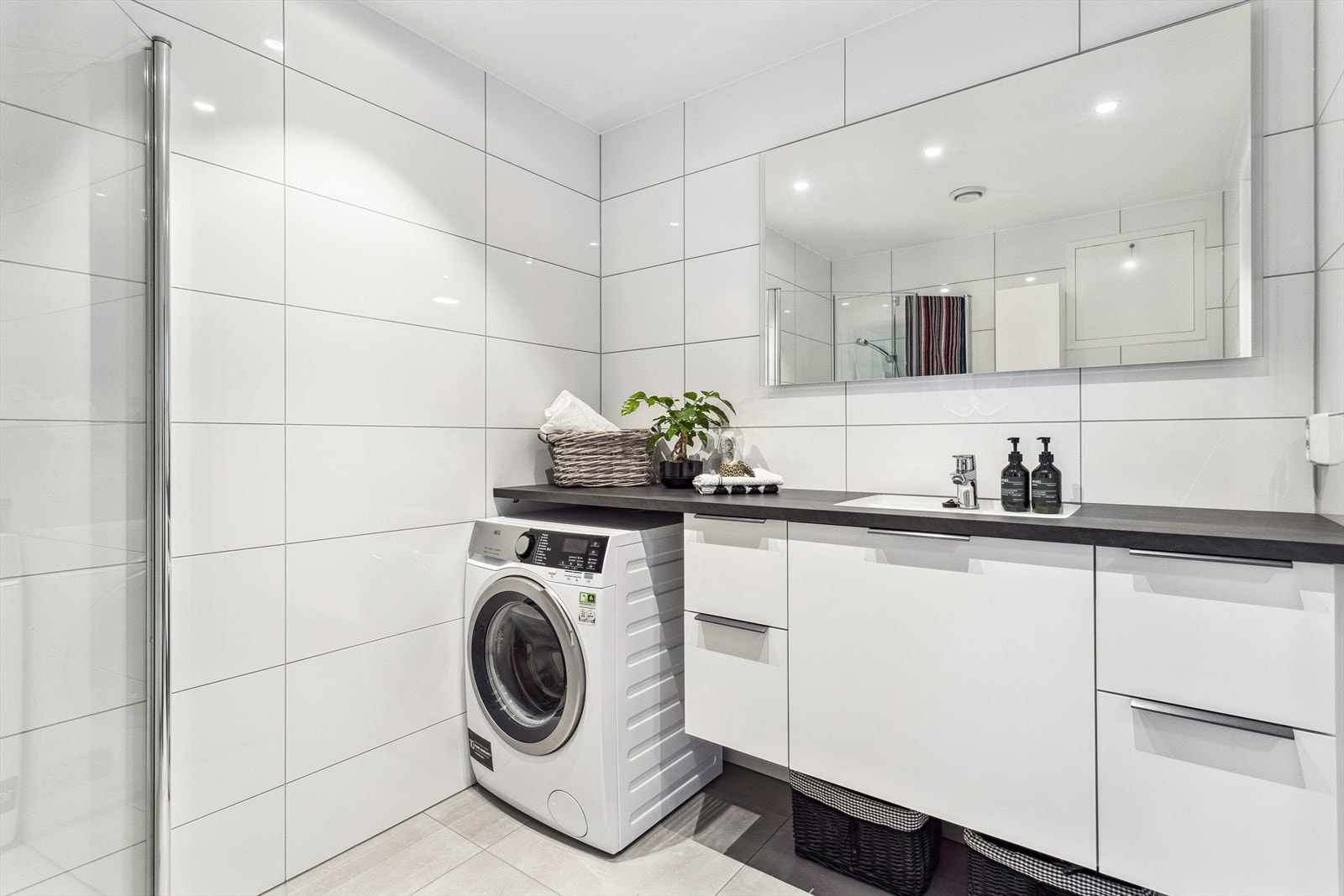 Badet er innredet med dusjvegger av glass og dusjgarnityr på vegg, vegghengt toalett, servant og opplegg for vaskemaskin. Galleribilde
