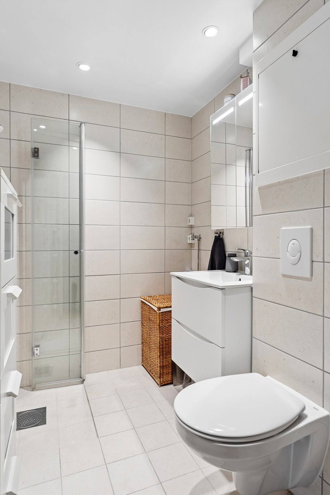 Badet har flislagt gulv med elektriske varmekabler og fliser på veggene. Innredningen inkluderer servantmøbel, vegghengt toalett og dusjhjørne med innfellbare glassdører. Det er opplegg for vaskemaskin Galleribilde