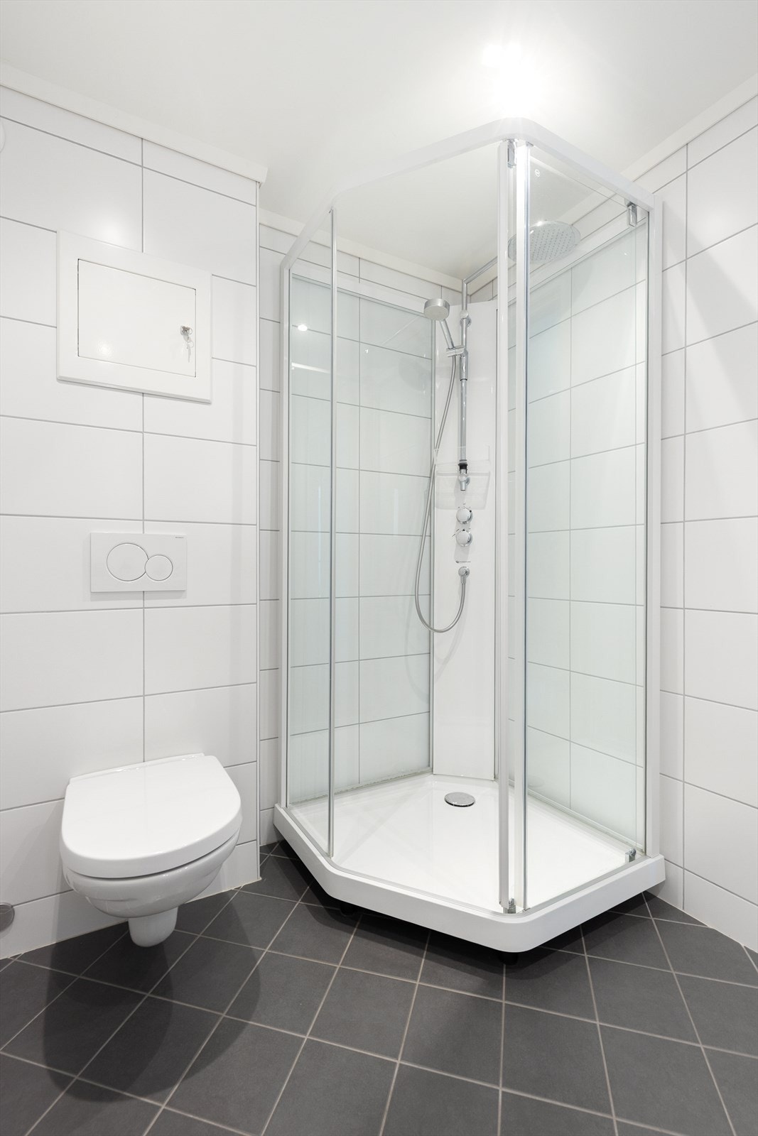 Badet har vegghengt toalett med innebygd sisterne, dusjkabinett og belysning med innfelte spotter i tak. Det er mekanisk avtrekk med kanal ut av badet. Galleribilde