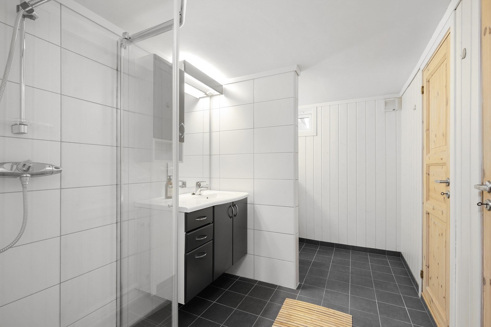 Badet har en romslig innredning i mørk utførelse med heldekkende servant, sideskap og speil med overlys og el-stikk. Badet er også innredet med åpen dusj med glassdører og veggmontert toalett. Galleribilde