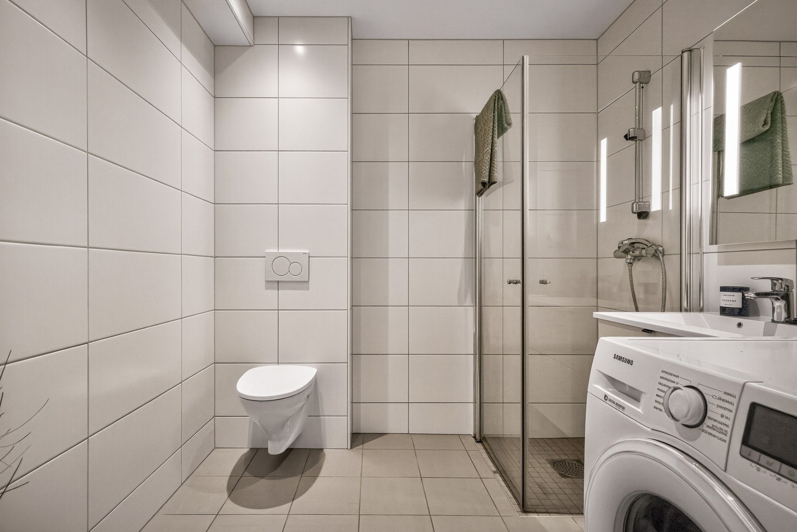 - Badet fremstår lyst og delikat, med flislagte overflater, vegghengt toalett, dusj med glassdører og moderne innredning - Galleribilde