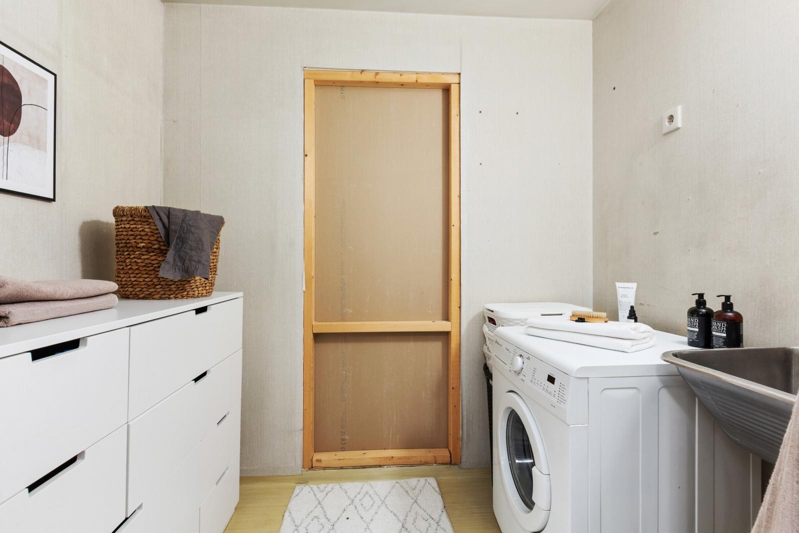 Vaskerom med opplegg for vaskemaskin og vaskekum. Vegger med lys tapet og belegg på gulv. Galleribilde