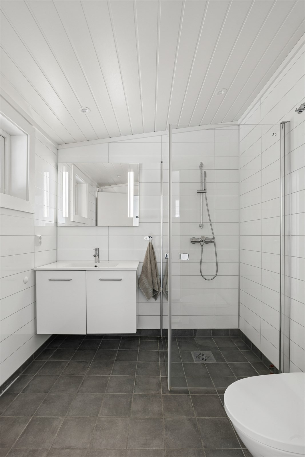 Delikat baderom med flislagt gulv og baderomsplater på vegg i lyse, tidløse farger. Badet er innredet med dusjhjørne med glassvegger, toalett og servant med innredning. Galleribilde