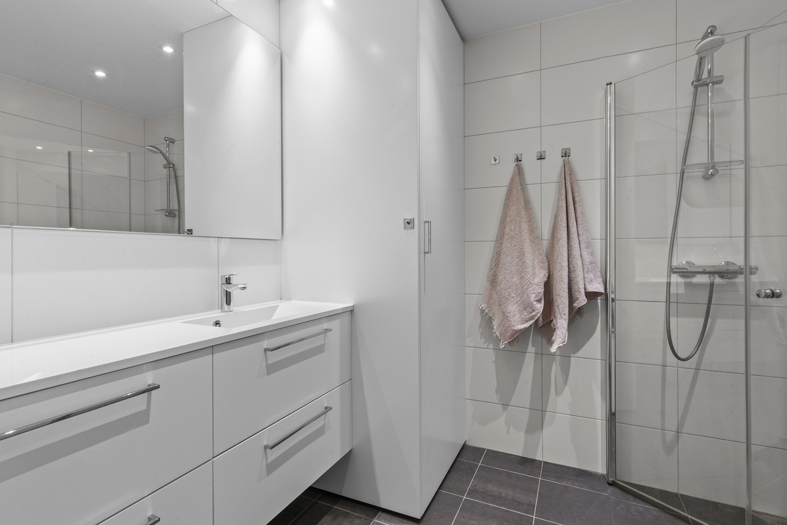 Dusjnisjen har praktiske glassvegger som sparer plass, mens innredningen består av servant med underskap, vegghengt toalett og integrert opplegg for vaskemaskin. Vannbåren gulvvarme og spotter i taket sørger for komfort og god belysning. Galleribilde
