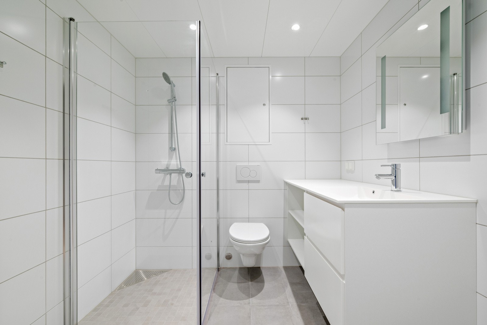 Badet har fliser på gulv, lekker baderomsinnredning med hvite profilerte fronter, vegghengt wc og opplegg for vaskemaskin Galleribilde