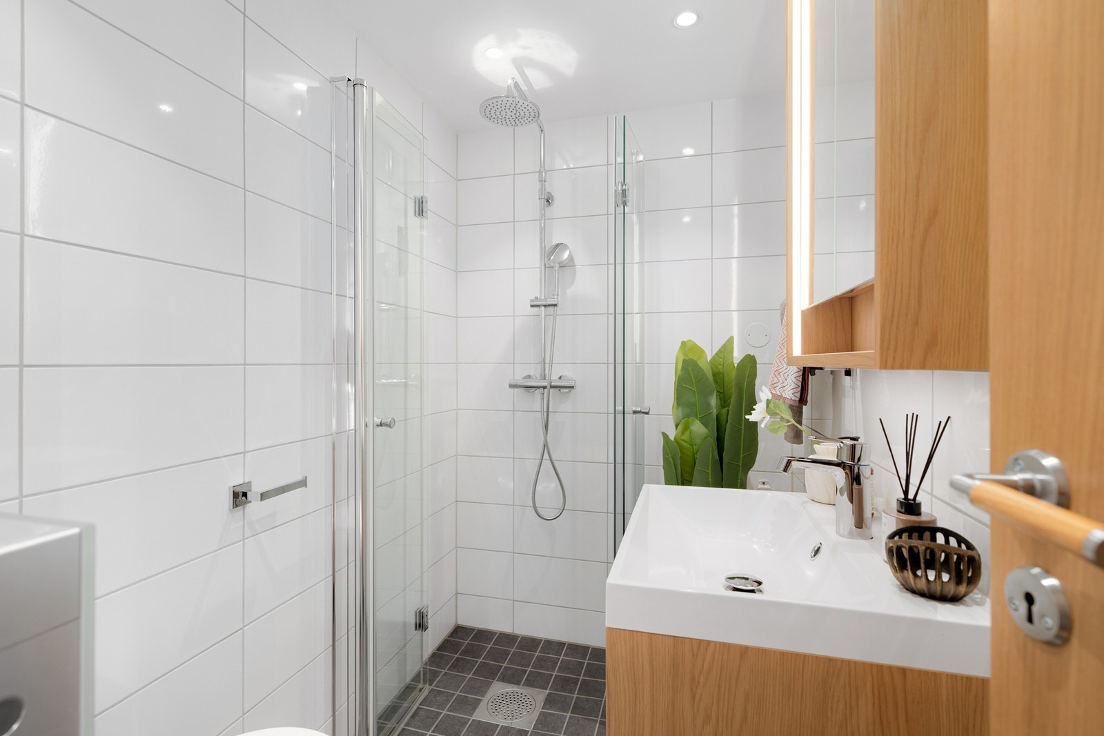 Baderommet har mørke fliser på gulv med gulvvarme, hvite fliser på veggene og downlights i himlingen. Galleribilde