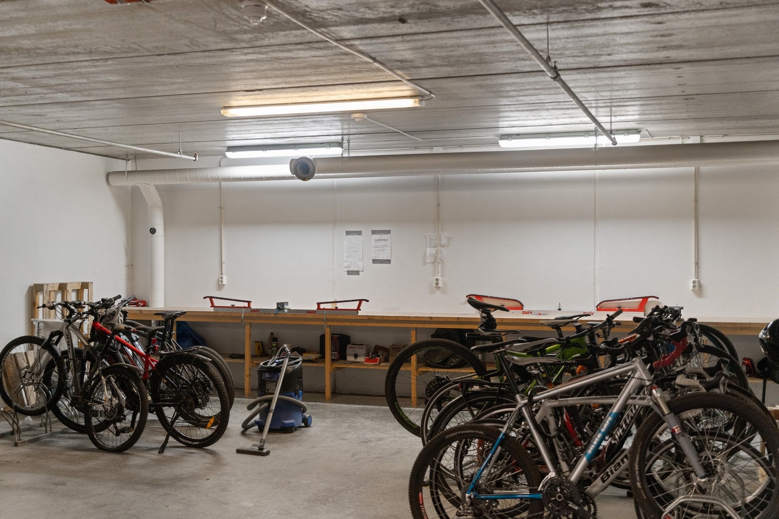 Felles sykkelparkering inne i garasjekjeller med stort arbeidsområde for sykkelreparasjoner. Felles høyttrykksspyler og industristøvsuger til sykkel og bilvask. Galleribilde