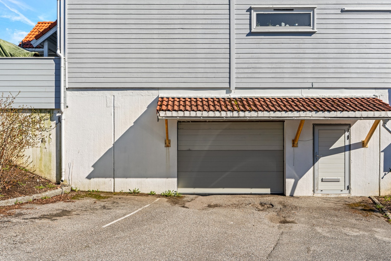 Garasje med tilhørende dør og overbygg, beliggende ved en gråmalt bygning. Asfaltert foran garasjen. Galleribilde