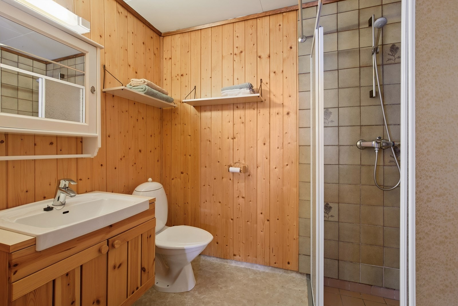 Badet i kjelleren er fra byggeår og er innredet med fliser på gulv og vegger i dusjsonen, panel på øvrige vegger og ellers vinylbelegg på gulvet. Badet er ellers utstyrt med dusjhjørne, toalett og baderomsinnredning med nedfelt servant. Galleribilde