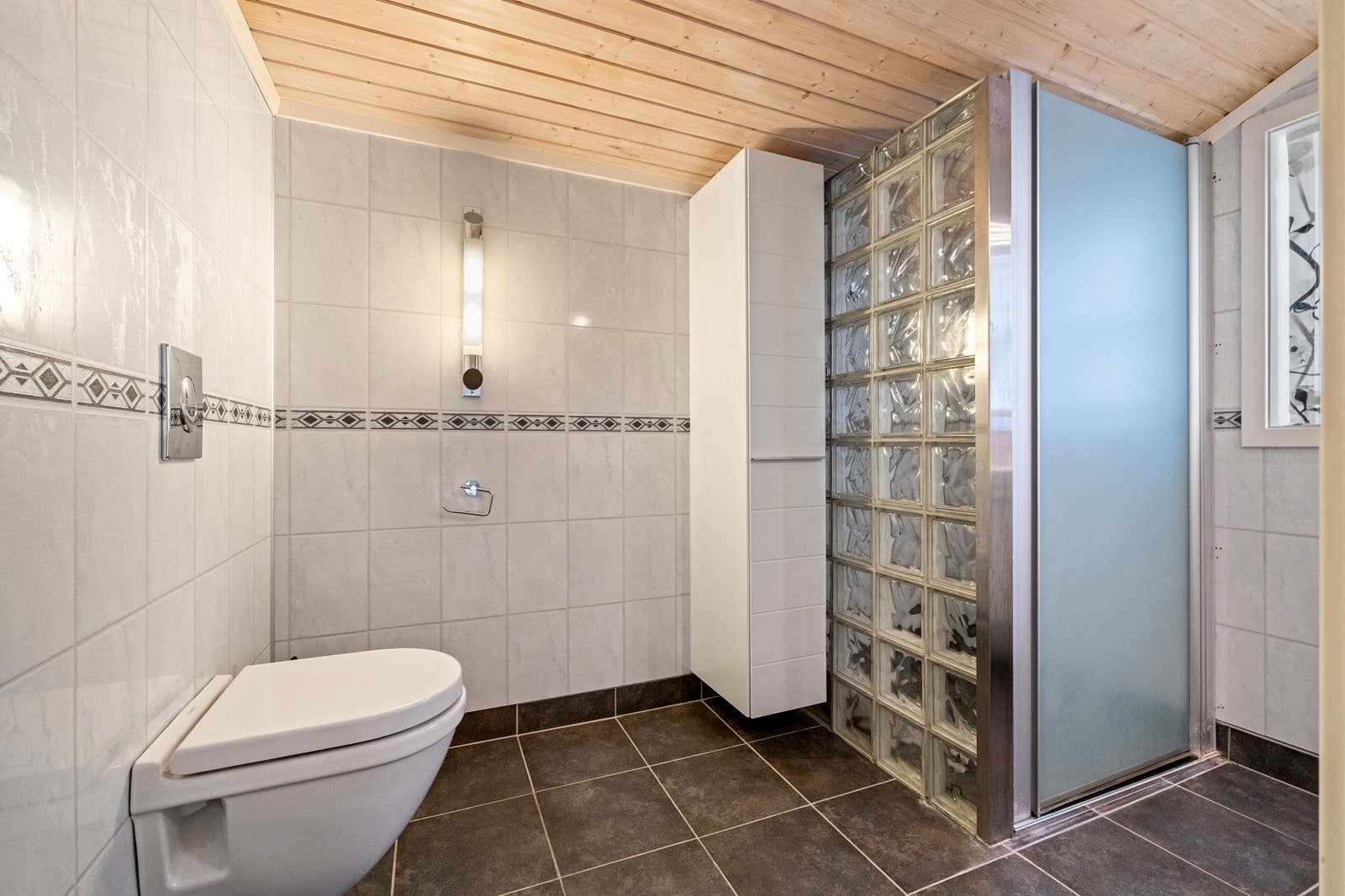 Badet er utstyrt med vegghengt wc, vegghengt høyskap og dushjørne med glassbyggerstein Galleribilde