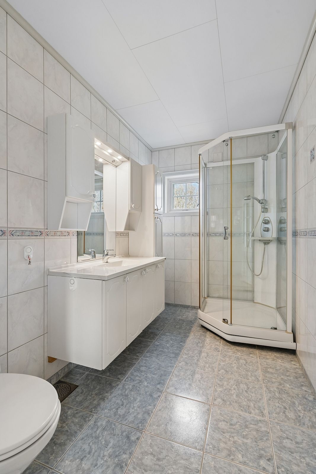 Badet har flislagte overflater og et dusjkabinett, samt ventilasjon gjennom en ventil i yttervegg. Galleribilde