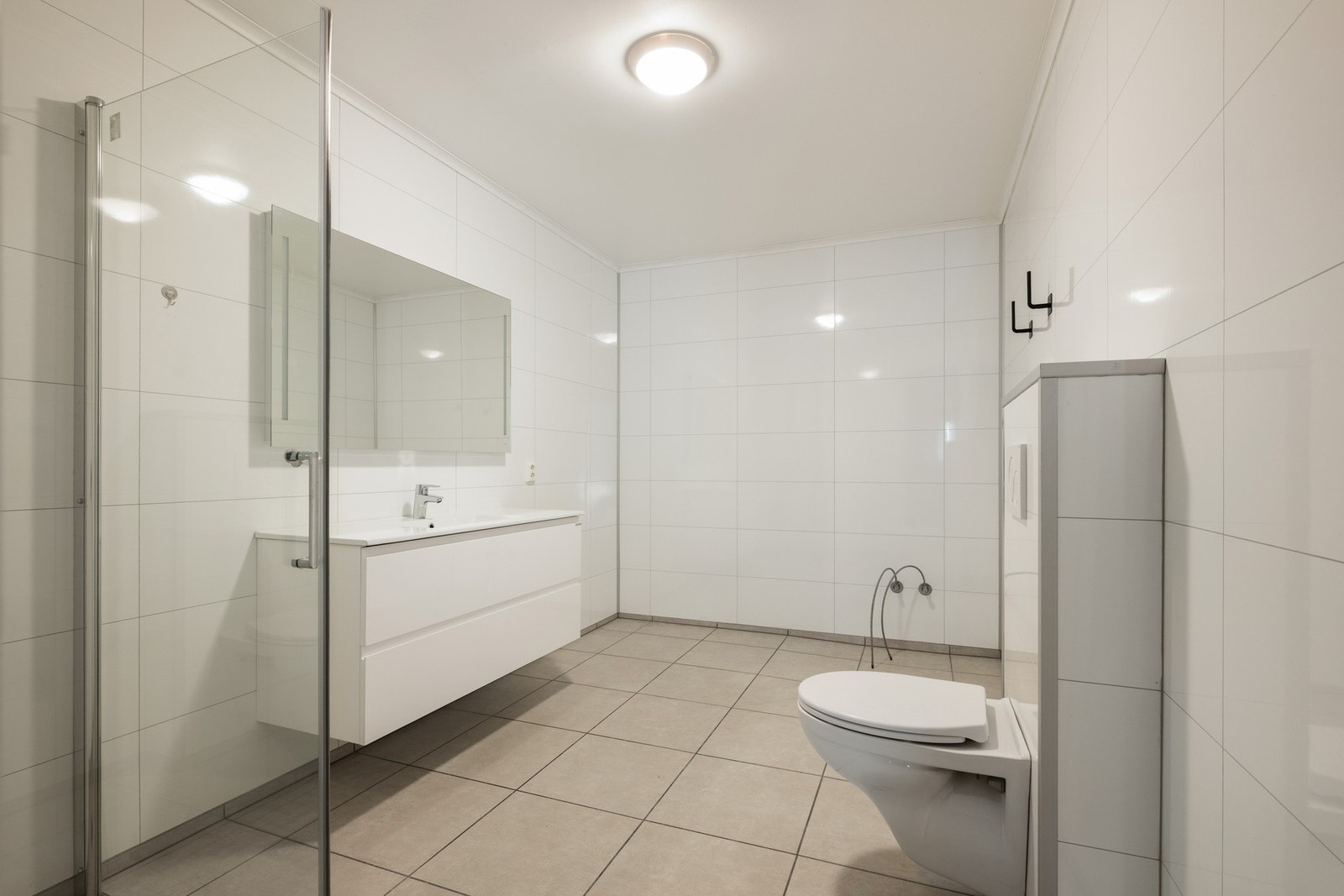 Badet har fliser på gulv og baderomsplater på vegger, vegghengt toalett, dusjhjørne og servant med speilskap. Galleribilde
