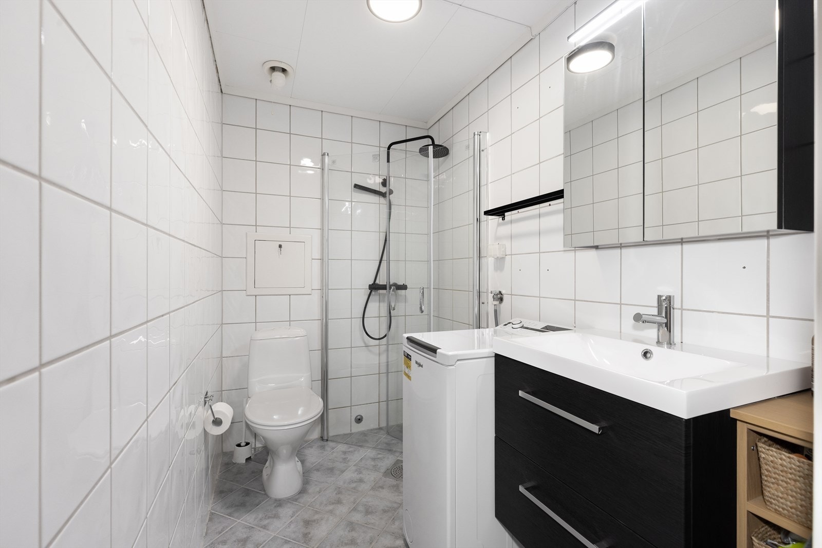 Pent flislagt baderom med varmekabler. Badet er utstyrt med gulvmontert toalett og dusjhjørne med glassdører, samt opplegg for vaskemaskin. Baderomsinnredning med skuffer, nedsenket servant ogspeilskap. Galleribilde