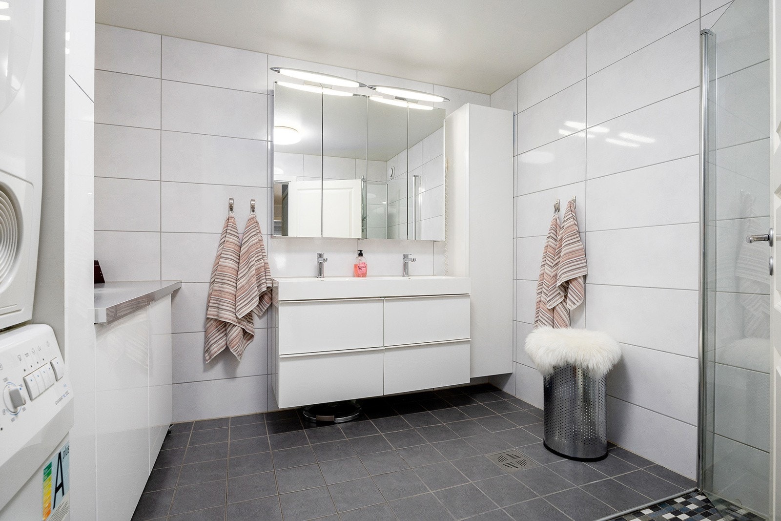 Moderne og gjennomtenkt bad med en elegant innredning bestående av dobbel servant med praktiske skuffer, stilrent speilskap med integrert belysning og et romslig dusjhjørne med hengslede glassvegger. Galleribilde