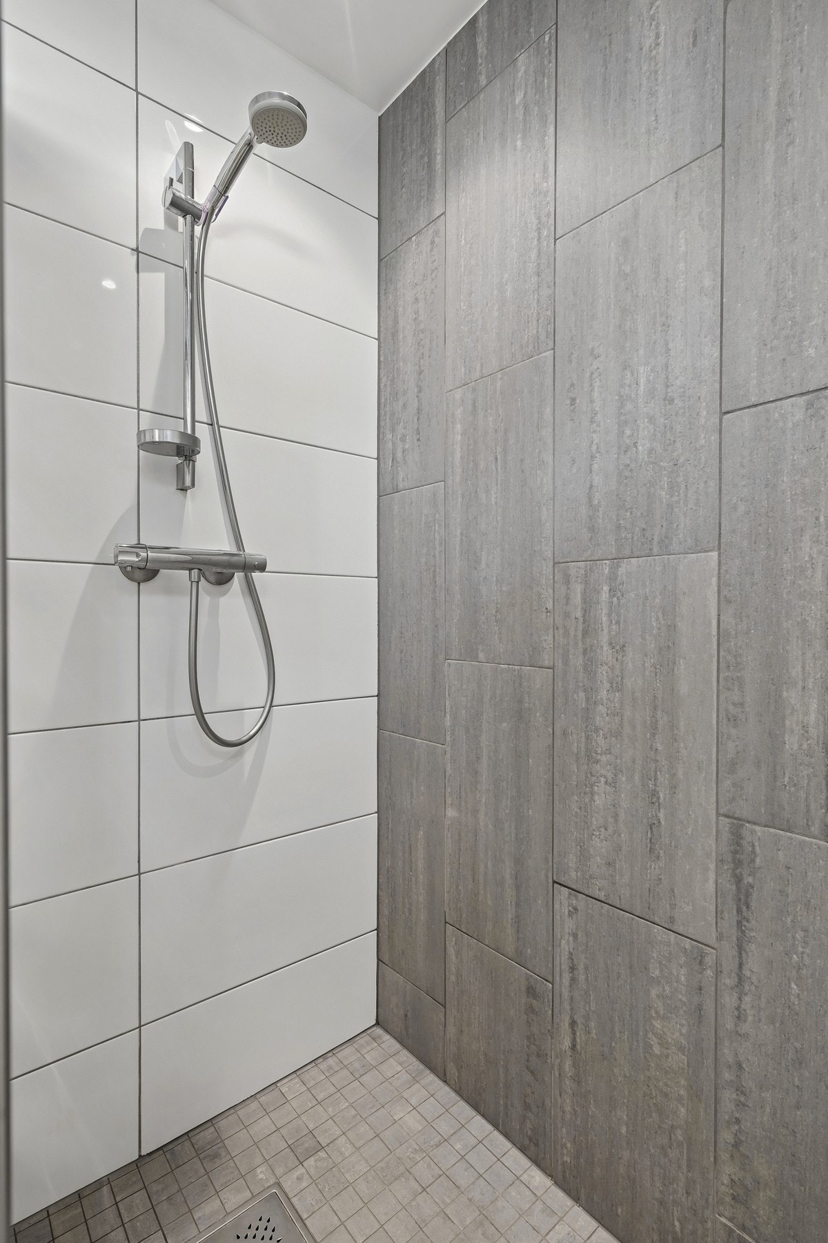 Badet er utstyrt med vegghengt toalett, dusjhjørne med hvite og grå fliser og vegghengt dusjarmatur. Galleribilde