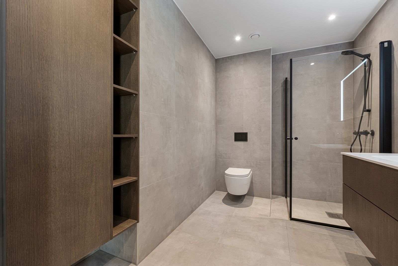 Badet er utstyrt med vegghengt toalett, dusjvegger med sorte detaljer og enkel servant med dobbel underskap Galleribilde