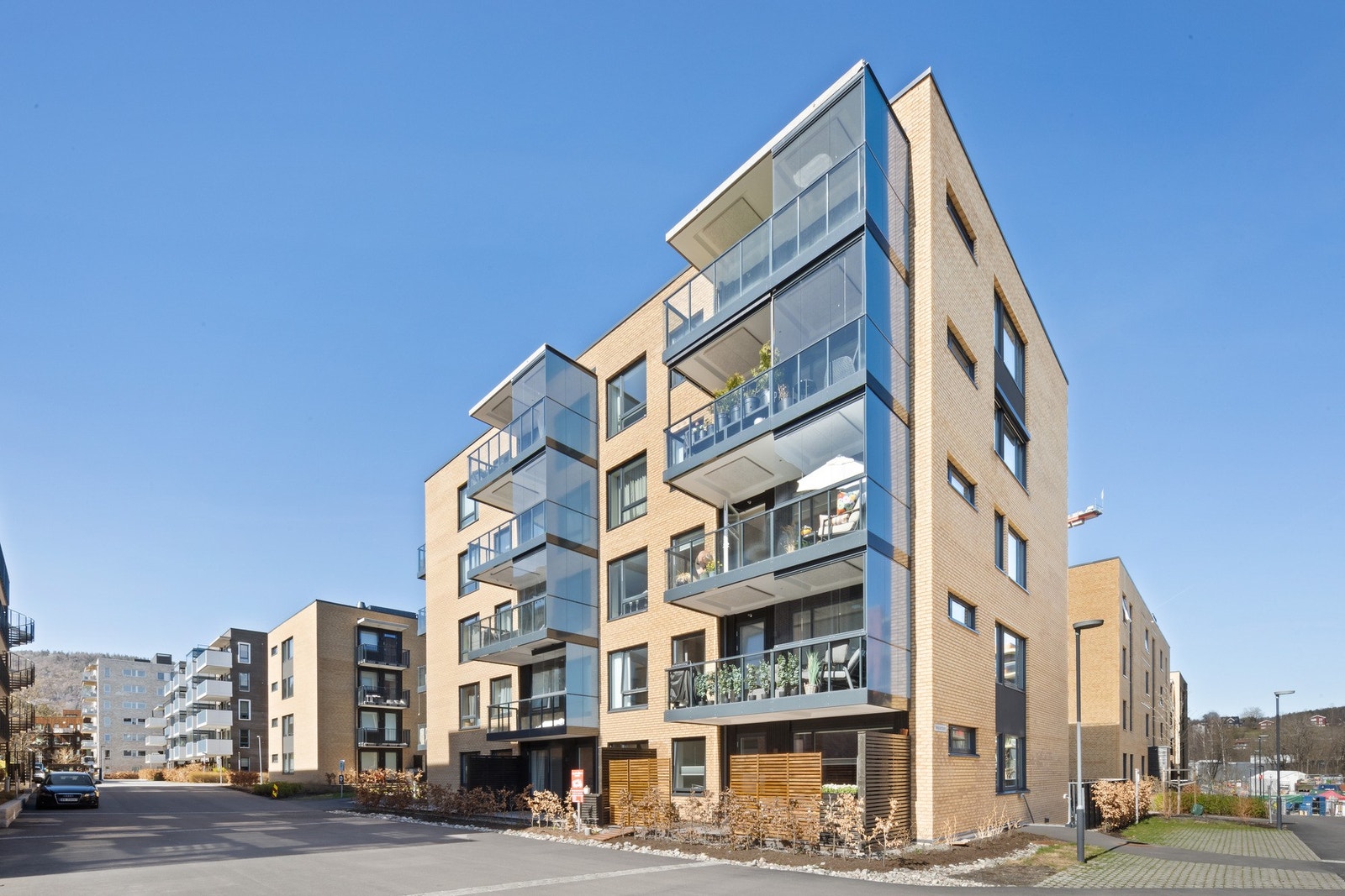 Moderne leilighetsbygg, fem etasjer, fasade i lys murstein og store glasspartier. Fellesområder med asfalterte gangveier og beplantning. Galleribilde