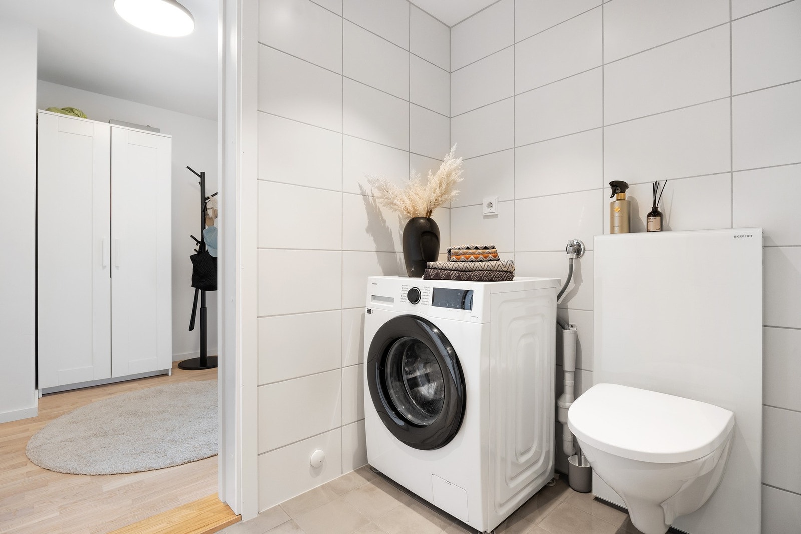 Lyst vaskerom med vaskemaskin og vegghengt toalett, tilknyttet gang med garderobeskap. Galleribilde