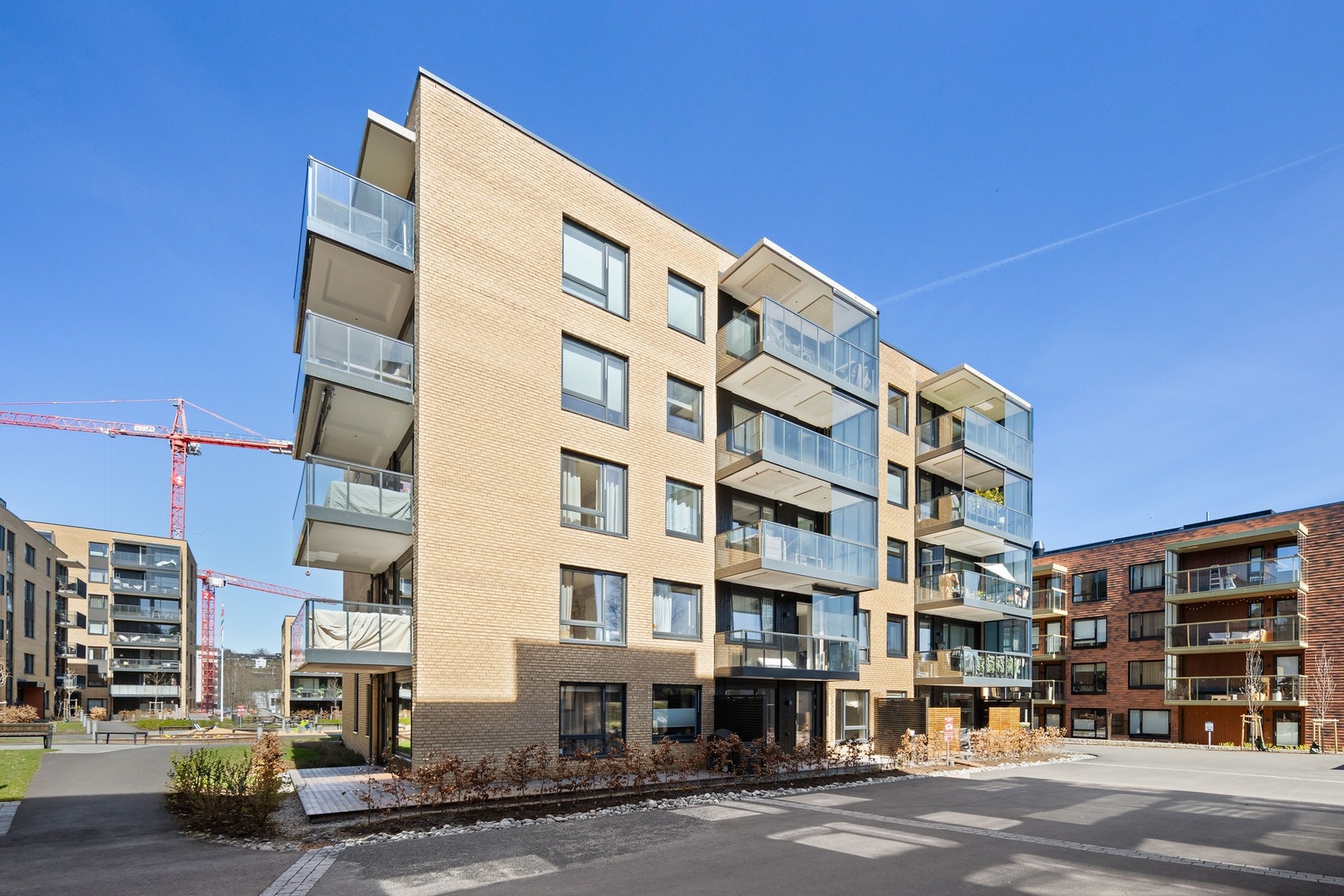 Bygget er en moderne boligblokk med fem etasjer, kledd i lys murstein og har balkonger med glassrekkverk. Galleribilde
