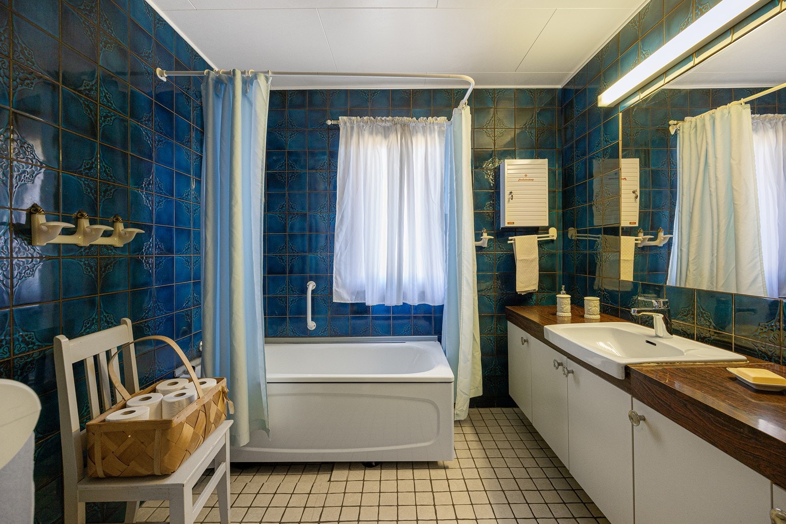Badet er romslig, med blå fliser på veggene og lyse fliser på gulv med elektrisk gulvvarme. Videre har badet et toalett og et badekar med dusjarmatur. Galleribilde