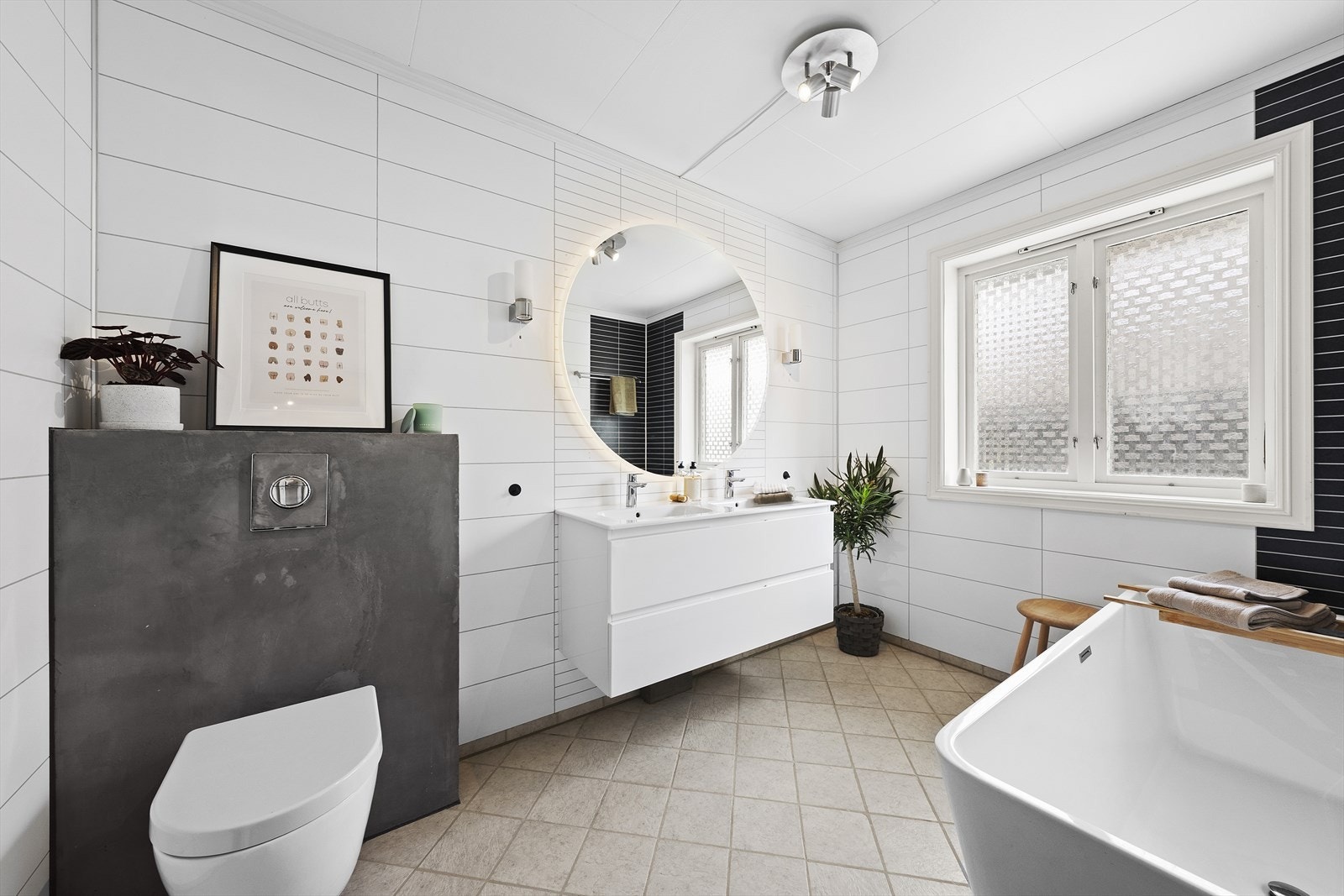 Badet er utstyrt med dobbel servant over skuffeinnredning, overliggende speil med belysning, badekar og vegghengt toalett Galleribilde