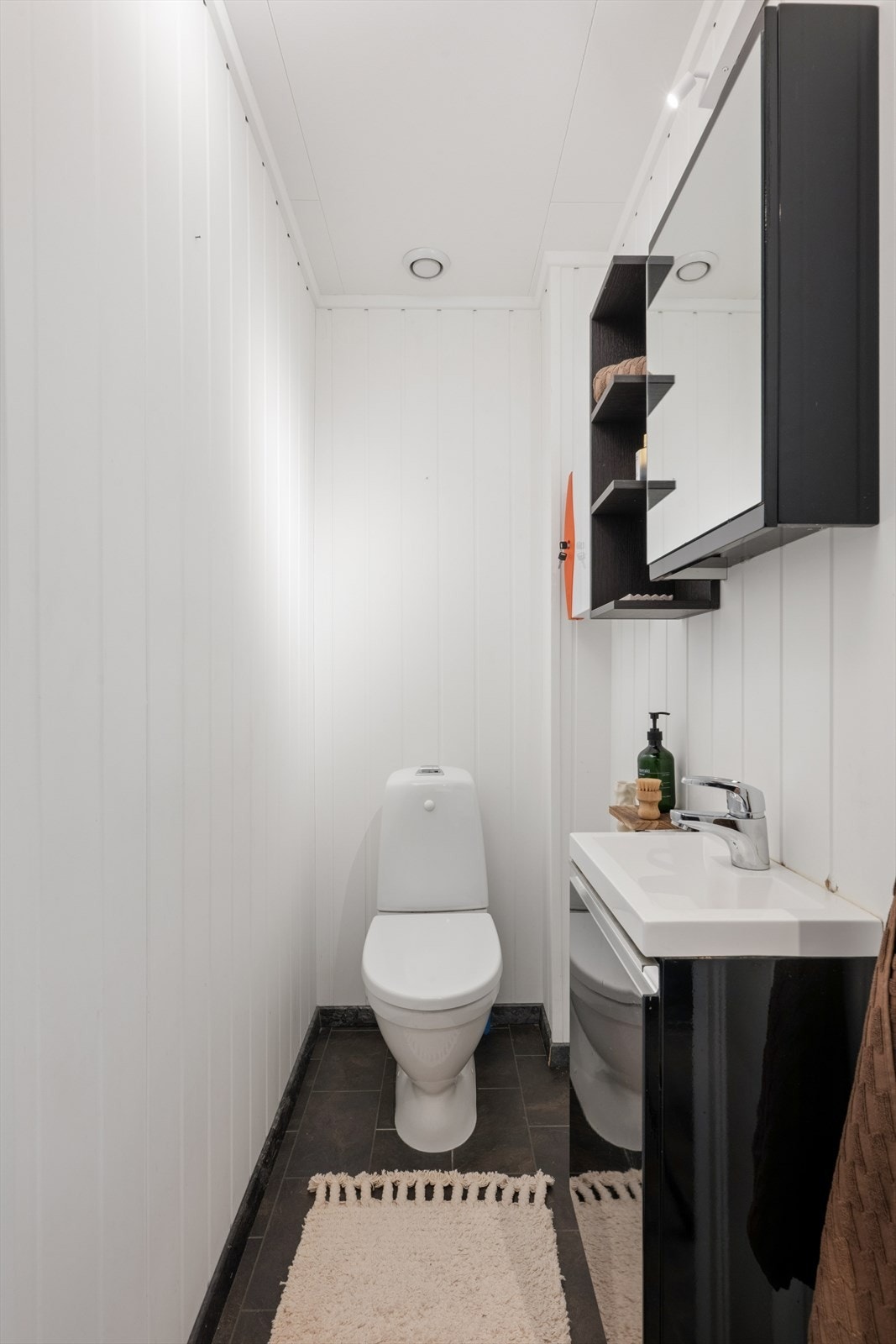 Wc-rommet ligger i tilknytning til entreen i første etasje og har flislaminat på gulvet, lysmalte vegger og et frittstående toalett. Rommet har også en heldekkende servant med mørke underskap, samt matchende vegghyller med speil. Galleribilde