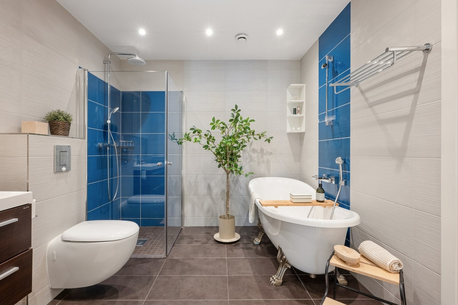 Badet har mørke fliser på gulvet og store, lyse fliser på veggene, samt gulvvarme, downlights, veggmontert toalett og et frittstående badekar. Galleribilde