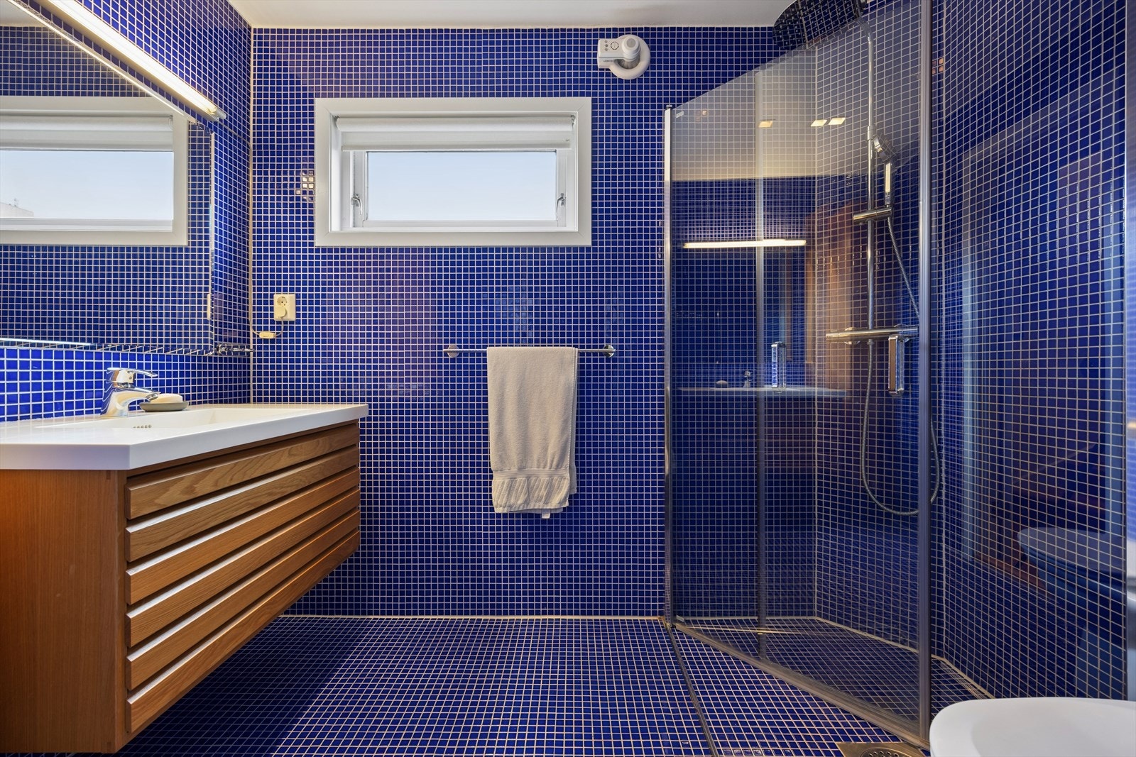 Helfliset bad med varme i gulv. Rommet er innredet med servant, toalett og dusjhjørne med glassvegger. Elektrisk styrt vifte for ventilasjon. Galleribilde