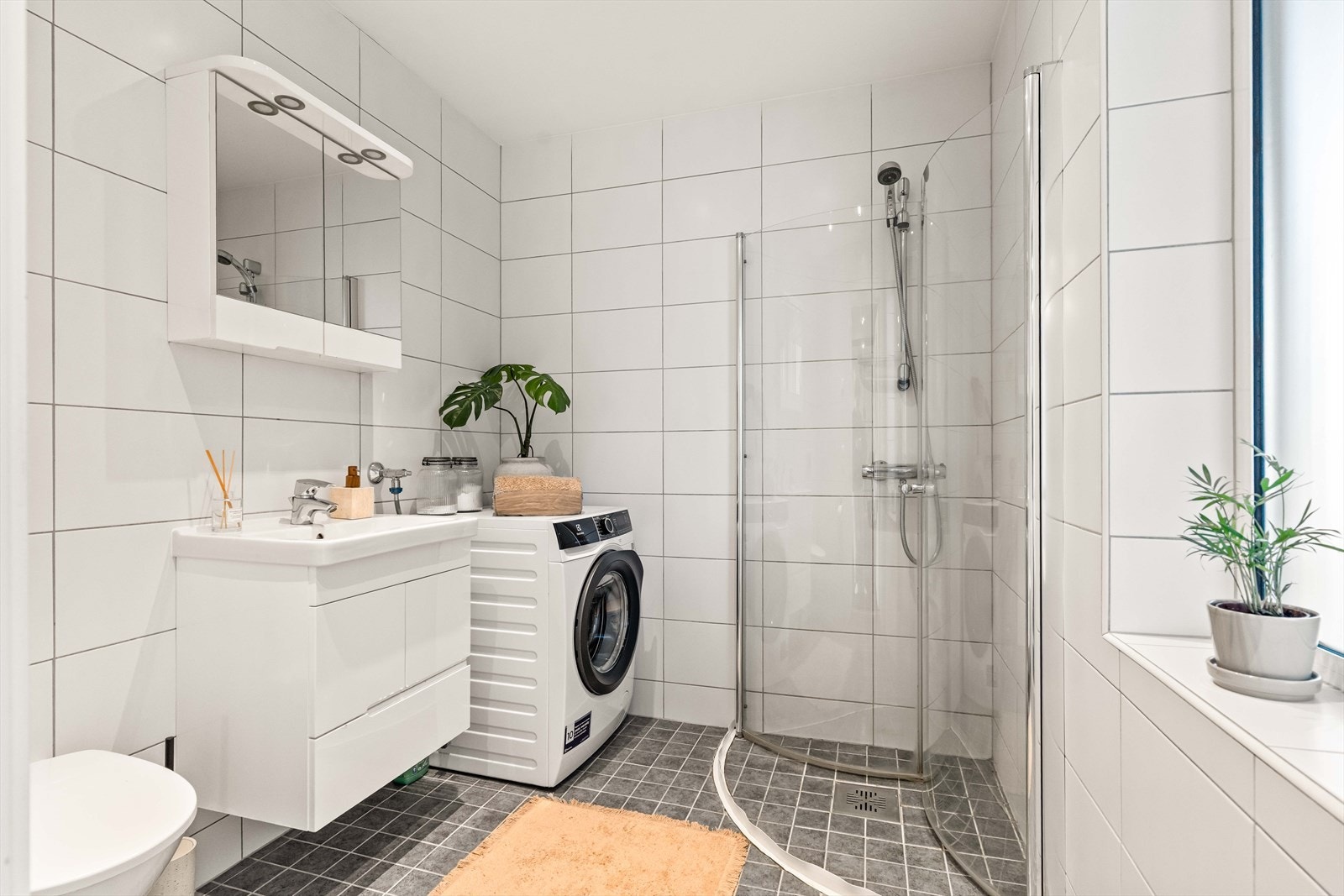 Moderne og delikat bad med fliser på gulv og vegg. Innredningen består av dusjhjørne med innfellbare dusjvegger, servant med skuffer, veggskap og speil med belysning, toalett og opplegg for vaskemaskin. Galleribilde