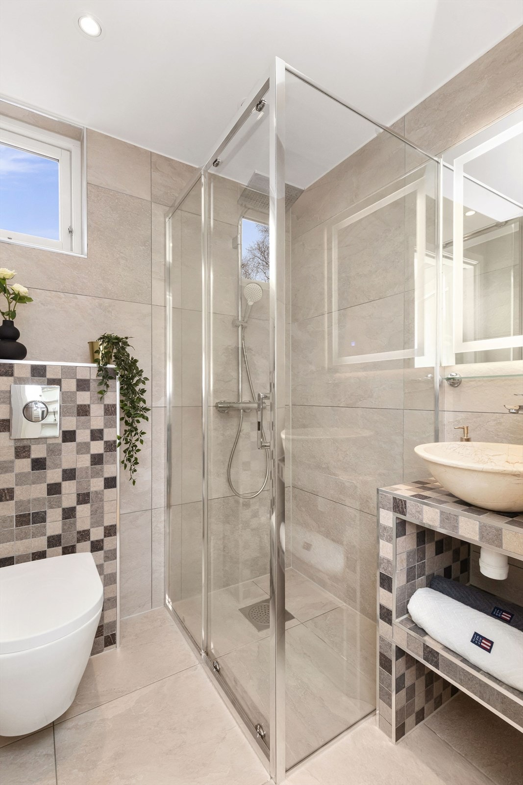 Badet ble oppusset ca. 2019 og fremstår innbydende med store fliser i lyse farger, romslig dusjsone med rainshower, veggmontert toalett og stilfull servant med speil og LED-lys. Galleribilde