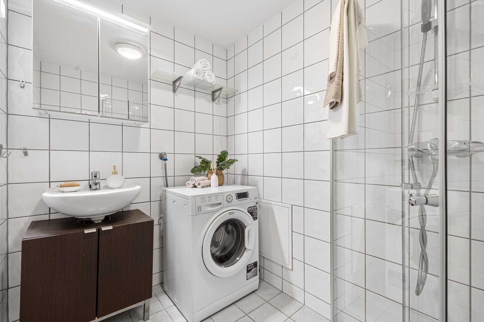 Baderommet inneholder innfellbare og plassbesparende dusjdører i glass, dusjbatteri og garnityr montert på vegg, kran og avløp for vaskemaskin og veggmontert WC. Rørskapet er lokalisert ved siden av vaskemaskinen. Galleribilde