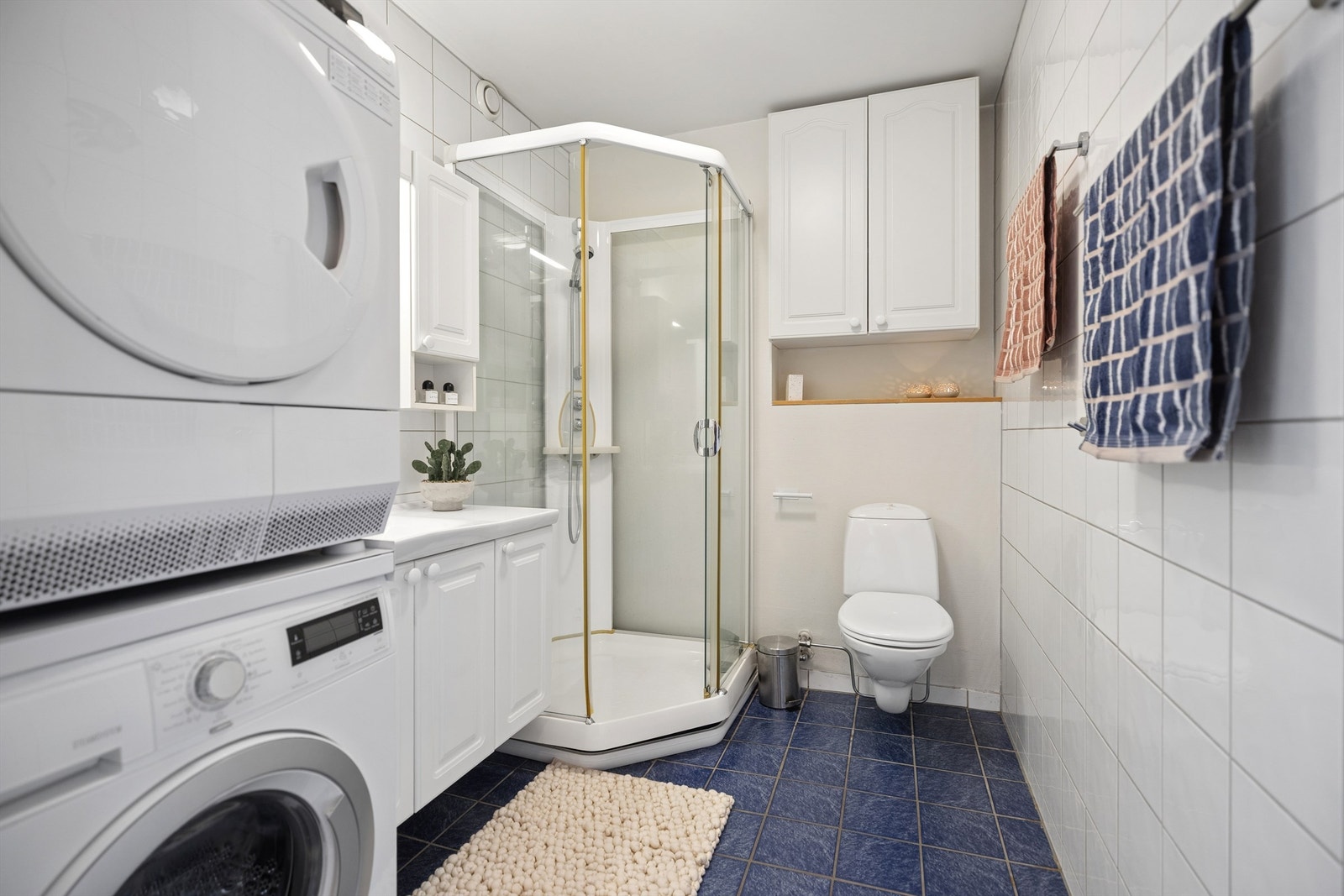 Lyst og delikat flislagt bad med behagelig gulvvarme som gir en lun start på dagen. Badet er innredet med servantskap, dusjkabinett og vegghengt toalett, samt praktisk plass for både vaskemaskin og tørketrommel. Galleribilde