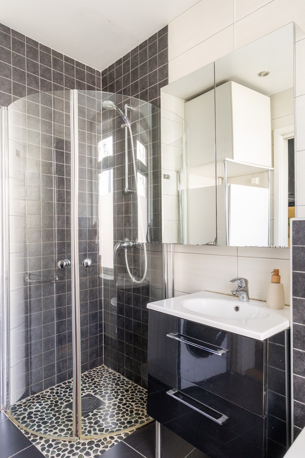 Badet er innredet med servant i skuffeseksjon og speilskap for oppbevaring. Videre er det veggmontert toalett og dusjhjørne med innfellbare glassdører. Galleribilde