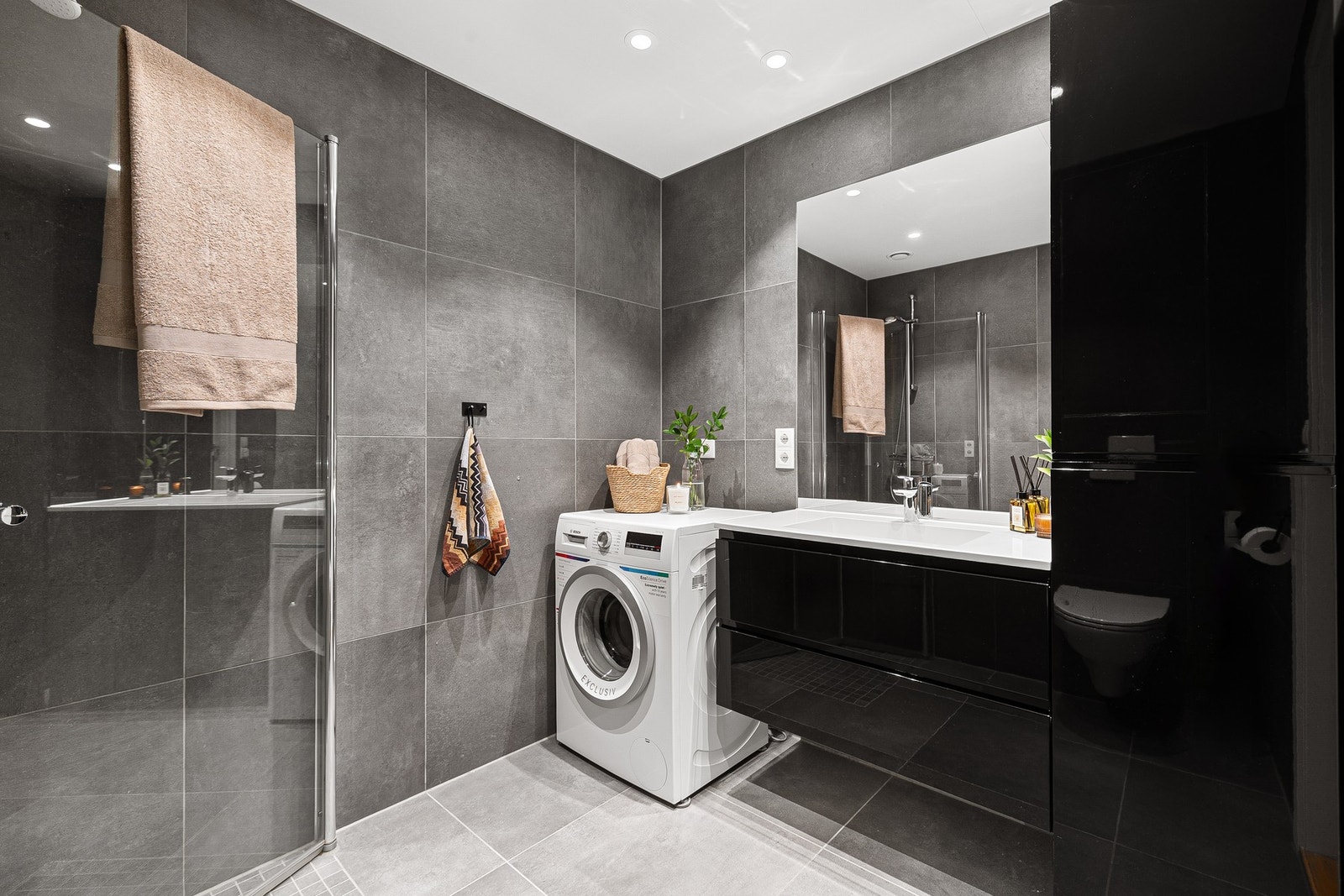 Badet inneholder innfellbare og plassbesparende dusjdører i glass, dusjbatteri og garnityr montert på vegg, veggmontert WC og kran og avløp til vaskemaskin. Spotter i himling. Galleribilde
