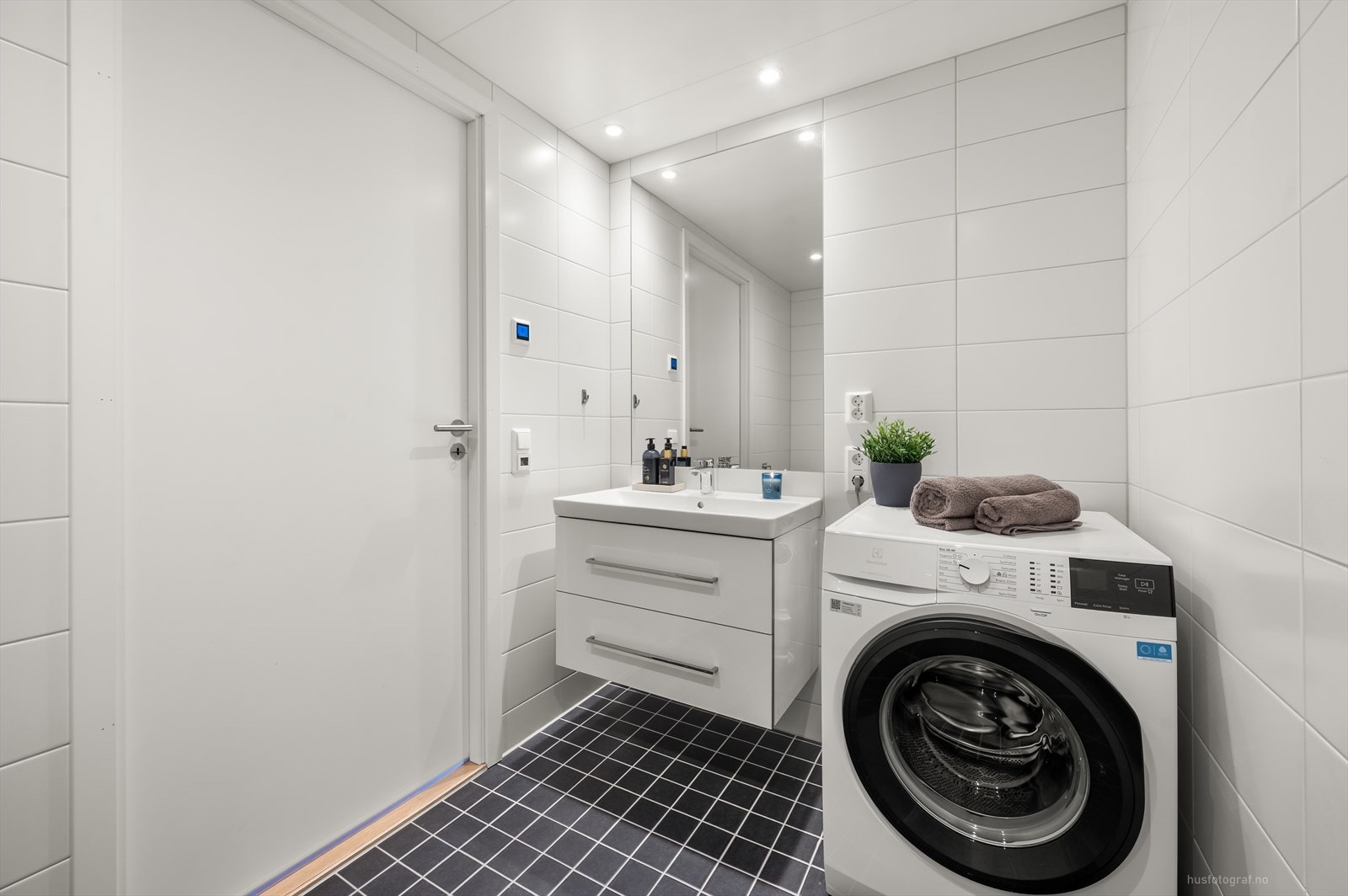 Badet i 1.etasje er flislagt med lyse fliser og innredningen består av servantinnredning med nedfelt servant med rom for oppbevaring i skuffer, speil med belysning, toalett og dusjnisje med dusjdør. Rommet har opplegg for vaskemaskin. Galleribilde