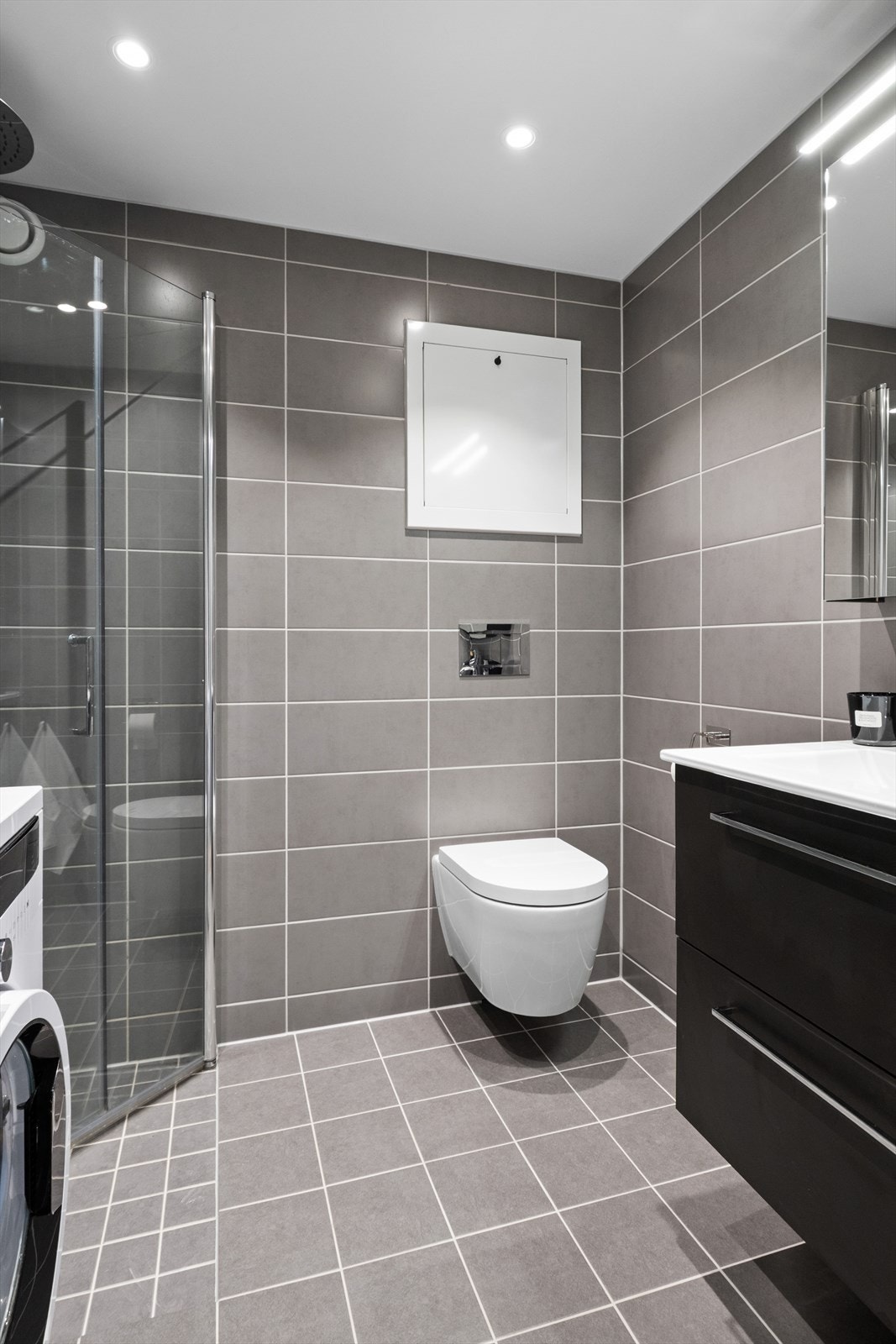 Badet er lekkert og har dusjvegger, vegghengt toalett, baderomsinnredning og opplegg til vaskemaskin Galleribilde