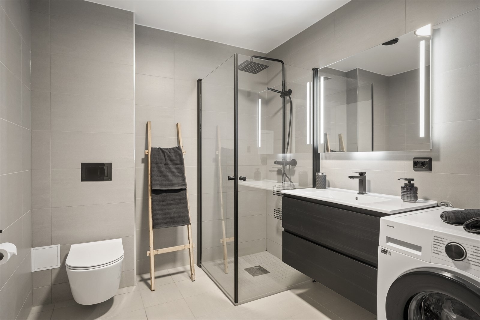 Badet er innredet mørke, glatte fronter, speil med lys, vegghengt toalett og dusjhjørne med glassdører Galleribilde